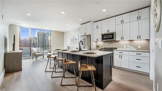 kitchen with a kitchen island with sink, a wall of windows, appliances with stainless steel finishes, white cabinetry, and light wood-type flooring