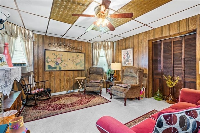 living room with a drop ceiling, wooden walls, ceiling fan, and carpet floors