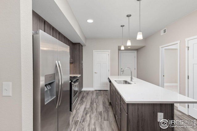 kitchen featuring light hardwood / wood-style flooring, appliances with stainless steel finishes, decorative light fixtures, dark brown cabinets, and a kitchen island with sink