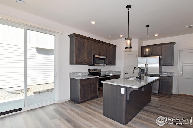 kitchen featuring stainless steel appliances, light stone counters, an island with sink, pendant lighting, and a kitchen bar