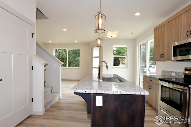 kitchen with stainless steel appliances, an island with sink, light stone counters, sink, and decorative light fixtures