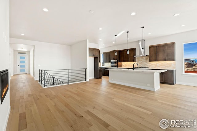 kitchen featuring backsplash, a center island with sink, hanging light fixtures, wall chimney exhaust hood, and light wood-type flooring