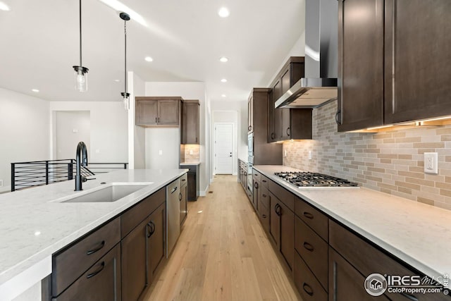 kitchen with sink, wall chimney exhaust hood, light stone counters, pendant lighting, and appliances with stainless steel finishes