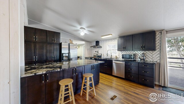 kitchen featuring wall chimney exhaust hood, a kitchen breakfast bar, stainless steel appliances, dark stone countertops, and vaulted ceiling