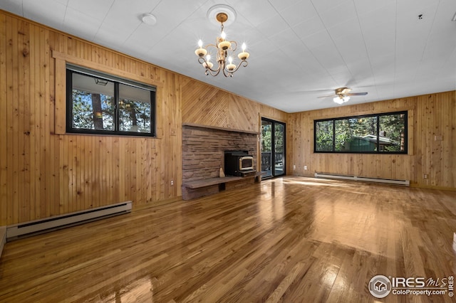 unfurnished living room with a wood stove, wood finished floors, wooden walls, a baseboard heating unit, and ceiling fan with notable chandelier
