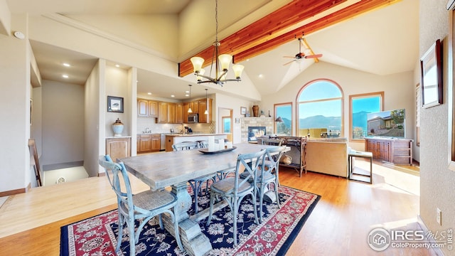 dining space featuring light hardwood / wood-style floors, a tile fireplace, beamed ceiling, ceiling fan with notable chandelier, and high vaulted ceiling