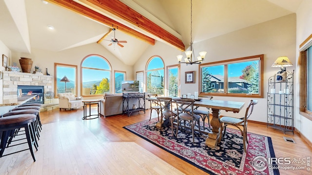 dining area with beamed ceiling, high vaulted ceiling, a fireplace, ceiling fan with notable chandelier, and light hardwood / wood-style floors