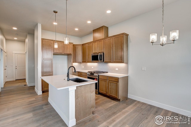 kitchen with an island with sink, appliances with stainless steel finishes, light wood-type flooring, sink, and decorative light fixtures