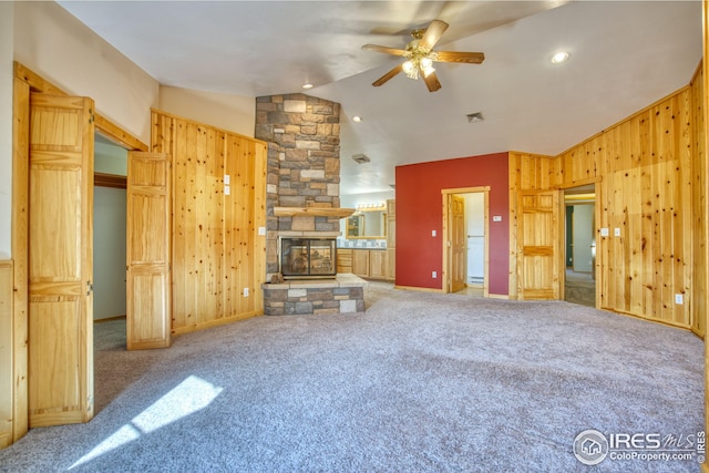 carpeted living room with a stone fireplace, ceiling fan, wooden walls, and vaulted ceiling