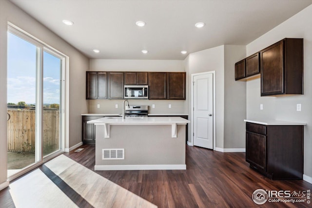 kitchen featuring dark hardwood / wood-style floors, dark brown cabinets, an island with sink, and appliances with stainless steel finishes