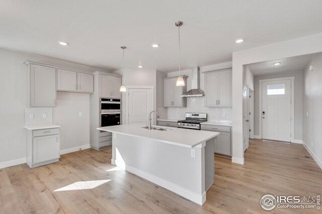 kitchen featuring a center island with sink, wall chimney range hood, sink, appliances with stainless steel finishes, and light hardwood / wood-style floors