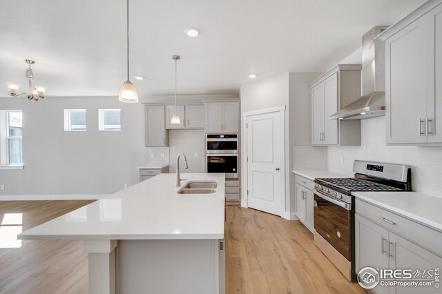 kitchen featuring sink, wall chimney range hood, an island with sink, decorative light fixtures, and appliances with stainless steel finishes