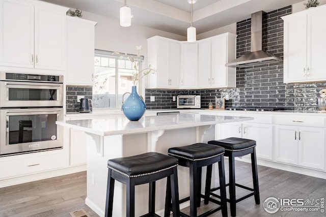 kitchen featuring white cabinetry, stainless steel double oven, pendant lighting, a kitchen island, and wall chimney exhaust hood