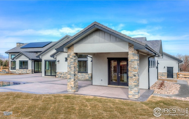 rear view of house with stucco siding, stone siding, french doors, a yard, and solar panels