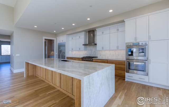 kitchen featuring light stone countertops, a sink, appliances with stainless steel finishes, wall chimney range hood, and light wood-type flooring