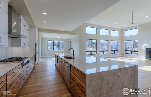 kitchen with wall chimney range hood, open floor plan, light wood-type flooring, stainless steel appliances, and a sink