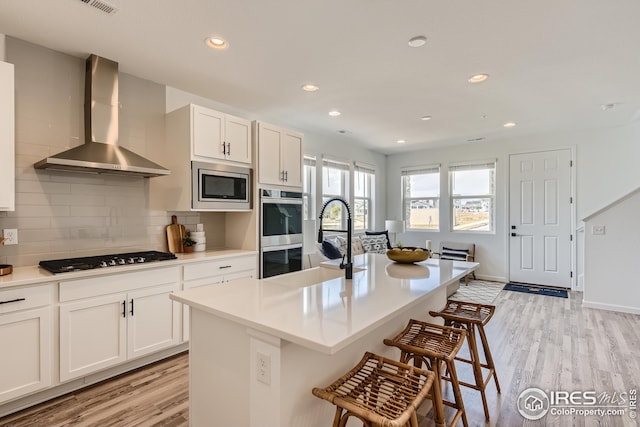 kitchen featuring white cabinetry, stainless steel appliances, wall chimney range hood, an island with sink, and light wood-type flooring