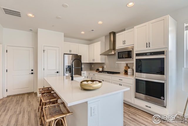 kitchen featuring a kitchen island with sink, a breakfast bar area, light hardwood / wood-style flooring, wall chimney exhaust hood, and stainless steel appliances