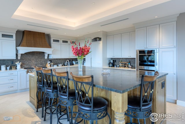 kitchen with a spacious island, custom exhaust hood, light tile patterned floors, a tray ceiling, and white cabinets