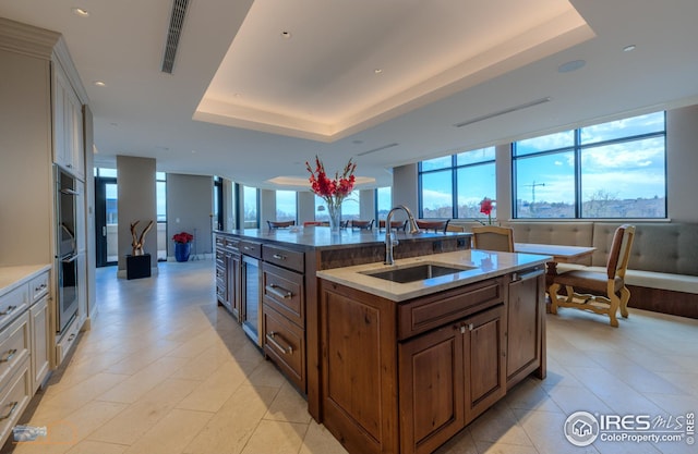 kitchen with sink, a tray ceiling, double oven, light stone countertops, and a kitchen island with sink