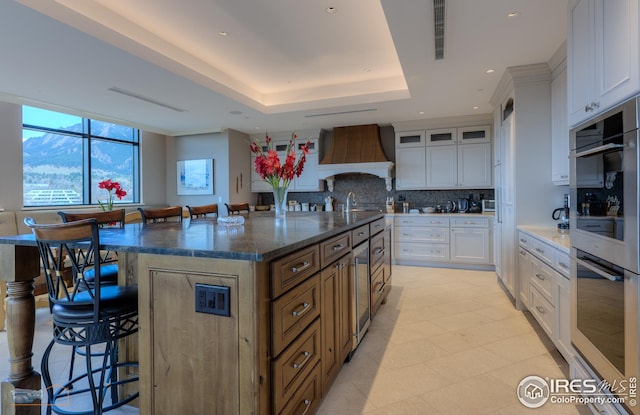 kitchen with a breakfast bar, tasteful backsplash, white cabinetry, a large island with sink, and custom range hood