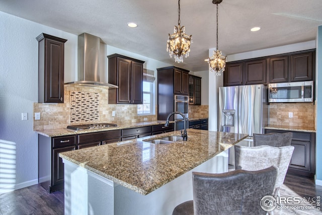 kitchen featuring wall chimney exhaust hood, stainless steel appliances, sink, decorative light fixtures, and an island with sink