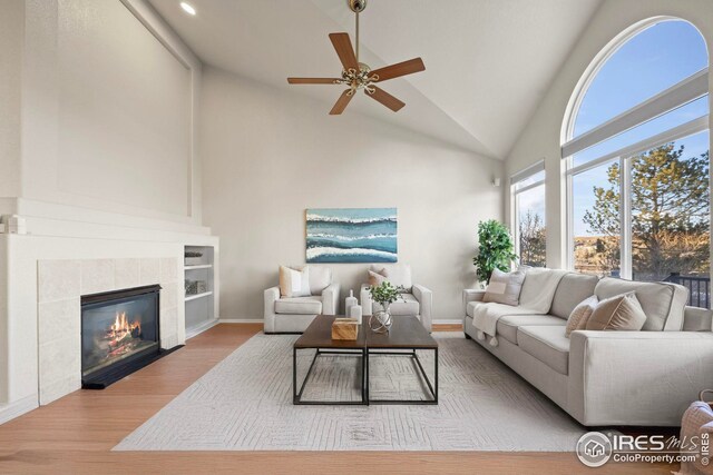 living area featuring high vaulted ceiling, baseboards, ceiling fan, a tiled fireplace, and wood finished floors
