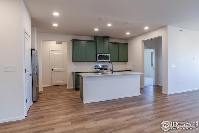 kitchen with appliances with stainless steel finishes, an island with sink, sink, green cabinets, and light hardwood / wood-style flooring