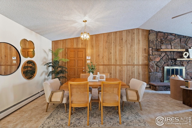 dining space featuring a baseboard radiator, lofted ceiling, a stone fireplace, wooden walls, and a chandelier
