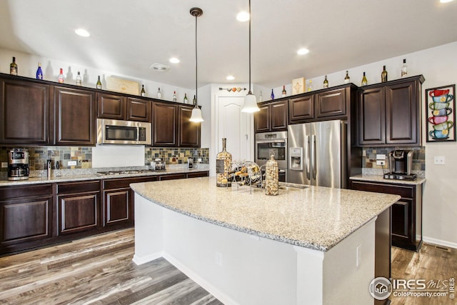 kitchen featuring appliances with stainless steel finishes, wood-type flooring, decorative backsplash, light stone counters, and a center island with sink