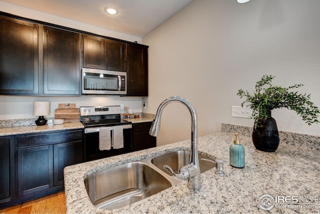 kitchen with vaulted ceiling, appliances with stainless steel finishes, sink, dark brown cabinetry, and light stone countertops