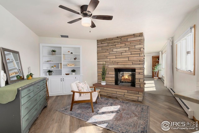 living room featuring dark wood finished floors, visible vents, baseboard heating, ceiling fan, and a stone fireplace