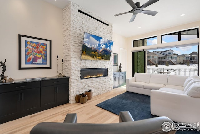 living room featuring ceiling fan, a stone fireplace, light wood finished floors, and a towering ceiling