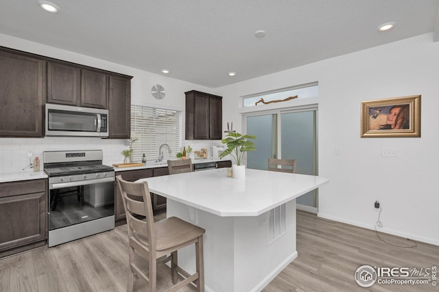 kitchen with dark brown cabinets, a breakfast bar, decorative backsplash, light wood-style flooring, and appliances with stainless steel finishes