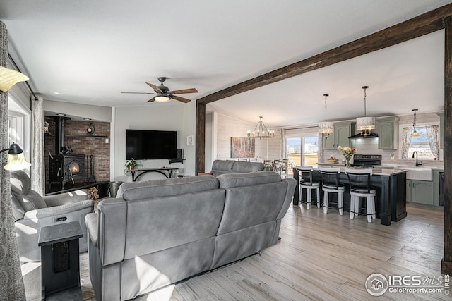 living room with ceiling fan with notable chandelier, sink, vaulted ceiling with beams, a wood stove, and light hardwood / wood-style flooring