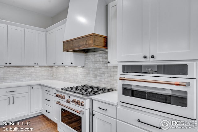 kitchen featuring white cabinetry, decorative backsplash, light hardwood / wood-style floors, custom range hood, and white appliances