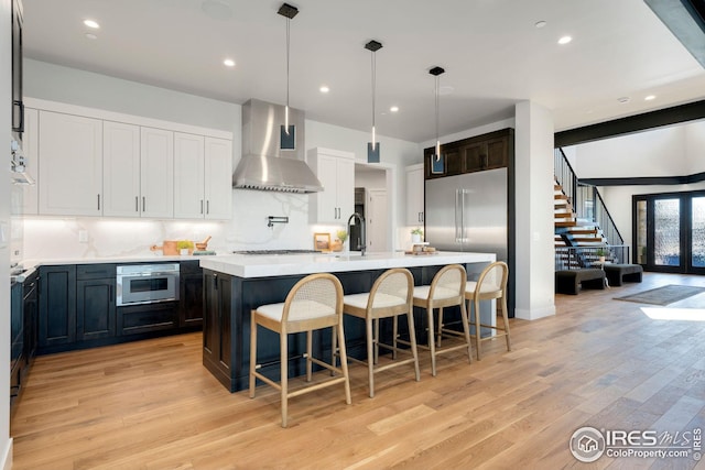 kitchen featuring stainless steel appliances, light wood-style floors, a center island with sink, wall chimney range hood, and light countertops