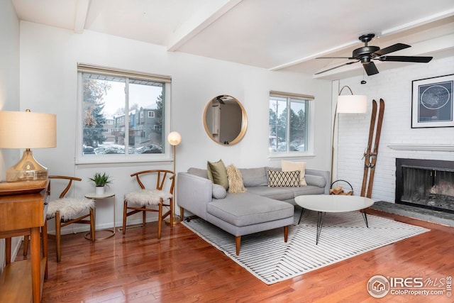 living room with a brick fireplace, beam ceiling, wood-type flooring, and ceiling fan