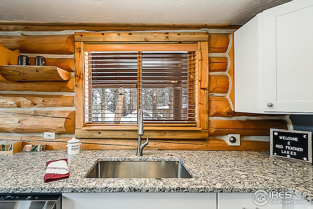 kitchen with light stone countertops, white cabinetry, rustic walls, a sink, and stainless steel dishwasher