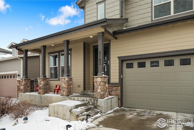 snow covered property entrance with a garage, a porch, concrete driveway, and stone siding