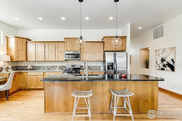 kitchen with light brown cabinets, hanging light fixtures, an island with sink, a sink, and appliances with stainless steel finishes