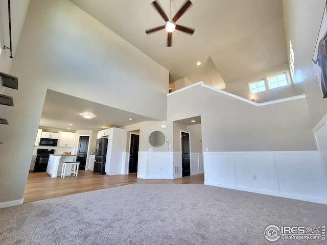 unfurnished living room featuring ceiling fan, high vaulted ceiling, wainscoting, and light carpet