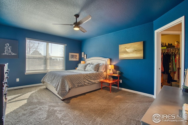 carpeted bedroom with a textured ceiling, a walk in closet, a ceiling fan, and baseboards