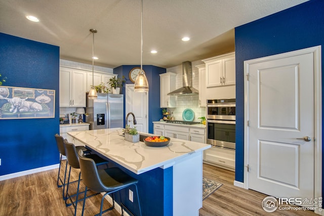 kitchen featuring light stone counters, stainless steel appliances, a kitchen island with sink, white cabinetry, and wall chimney exhaust hood