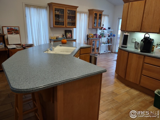 kitchen with brown cabinetry, an island with sink, vaulted ceiling, light wood-style floors, and a sink