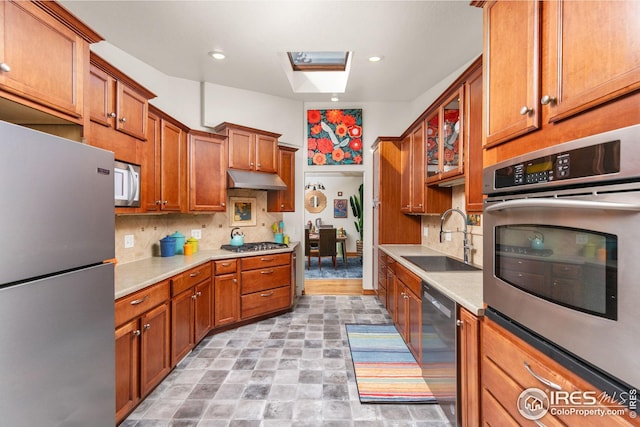 kitchen featuring under cabinet range hood, stainless steel appliances, a skylight, a sink, and brown cabinets