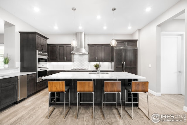 kitchen featuring a sink, tasteful backsplash, appliances with stainless steel finishes, light wood-type flooring, and wall chimney range hood