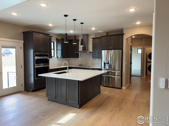 kitchen with arched walkways, a sink, stacked washer and dryer, appliances with stainless steel finishes, and wall chimney range hood