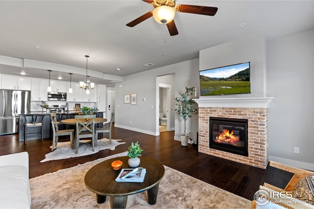 living area with baseboards, a fireplace, dark wood-style flooring, and ceiling fan with notable chandelier