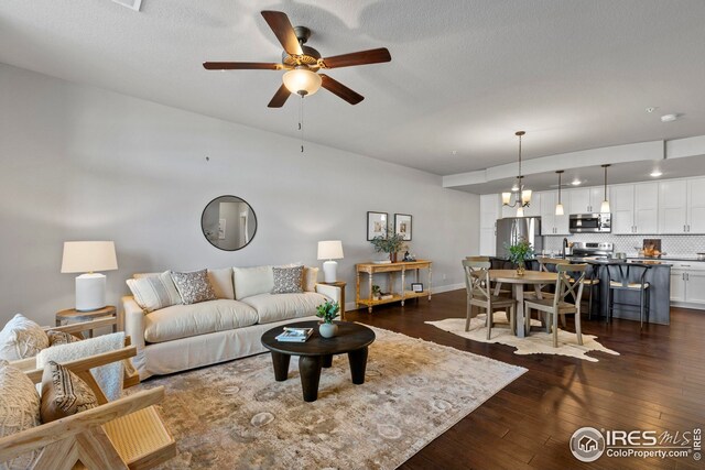 living room with a textured ceiling, ceiling fan with notable chandelier, dark wood-style flooring, and baseboards